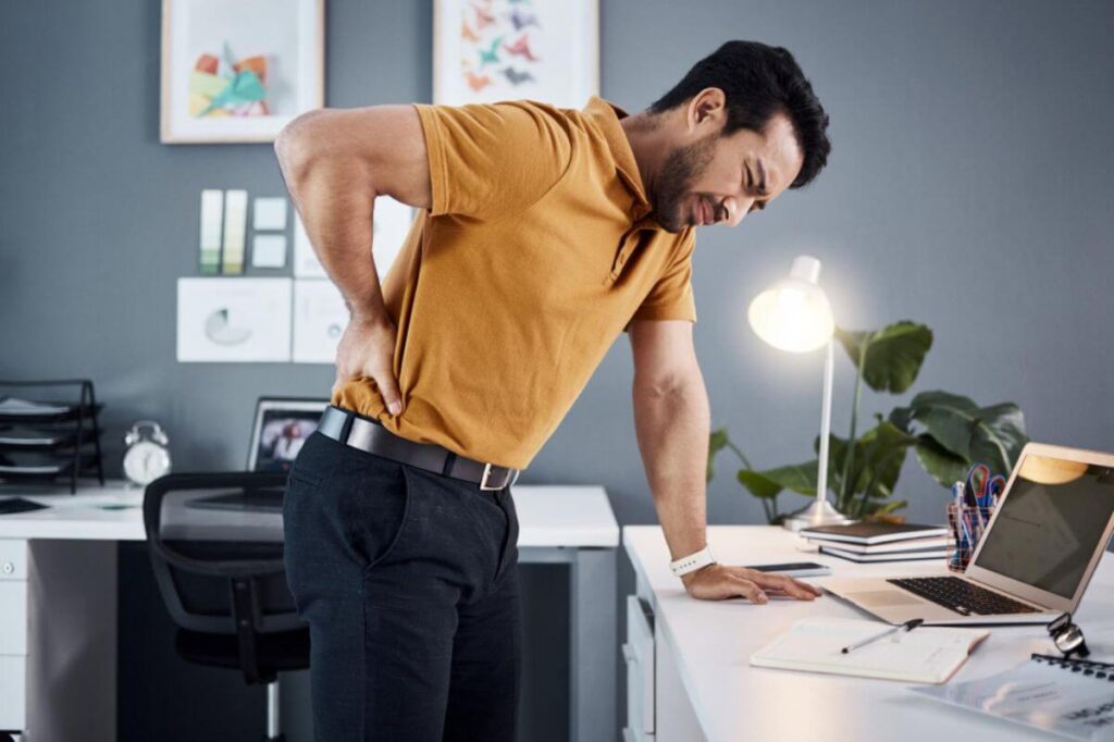Office worker holding his lower back from pain while standing beside his desk
